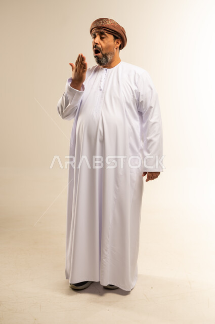 الإحساس بالتعب والإرهاق، ضرورة أخذ قسط من الراحة، التثاؤب والخمول والحاجة إلى النوم، بورتريه لرجل عربي خليجي عماني يرتدي الدشداشة والعمامة يضع يده أمام فمه بإيماءات النعاس والكسل، صورة لكامل الجسم، خلفية بيضاء