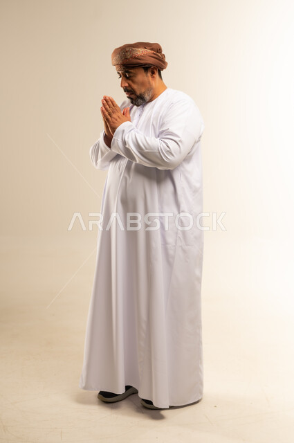 الإجتهاد في العبادة والحفاظ على الطاعات، الإكثار من الدعاء في شهر رمضان، بورتريه من الجانب لرجل عربي خليجي عماني يرتدي الدشداشة والعمامة يرفع يديه بإيماءات التوسل والتضرع، صورة الجسم بالطول الكامل، خلفية بيضاء