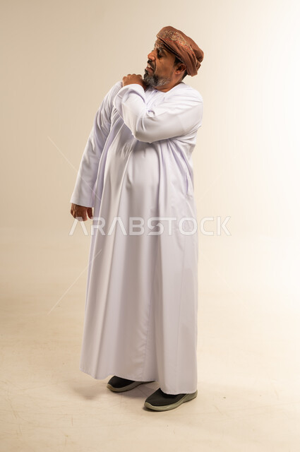 الأوجاع والآلام العصبية والعضلية، طلب المساعده والحاجه للعلاج، التهاب العضلات والمفاصل، بورتريه لرجل عربي خليجي عماني يرتدي الدشداشة والعمامة يضع يده على كتفه بايماءات الألم والتعب والأرهاق، حالة اسعافية طارئة، صورة الجسم الطول الكامل, خلفية بيضاء