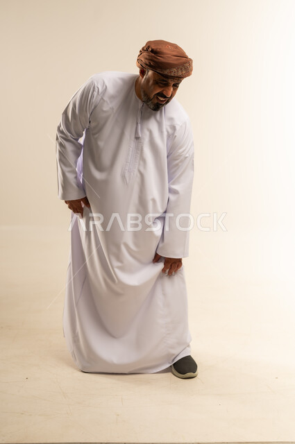 التهاب العضلات والمفاصل، الأوجاع والآلام العصبية والعضلية، طلب المساعده والحاجه للعلاج، بورتريه لرجل عربي خليجي عماني يرتدي الدشداشة والعمامة يضع يده على ركبته بايماءات الألم والتعب والأرهاق، حالة اسعافية طارئة،  صورة الجسم الطول الكامل, خلفية بيضاء