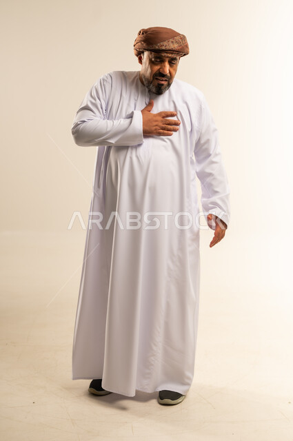 آلام الذبحة الصدرية، التعرض لنوبات قلبية مفاجئة، الإحساس بوخزة في الصدر والقلب، بورتريه لرجل عربي خليجي عماني يرتدي الدشداشة العمامة يضع يده على قلبه بإيماءات التوجع والألم، صورة الجسم بالطول الكامل، خلفية بيضاء