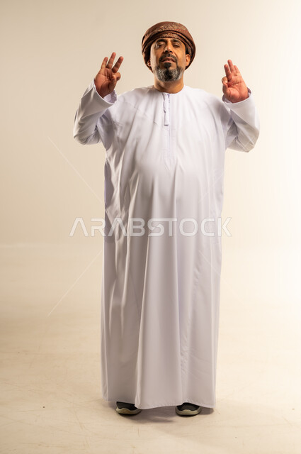 الشعور بالاعجاب والقبول، بورتريه لرجل عربي خليجي عماني يرتدي الدشداشة والعمامة يقف بعينين مغمضة يرفع يديه للأعلى بإيماءات الجودة، تعابير الرضا والسرور، صورة الجسم بالطول الكامل، خلفية بيضاء