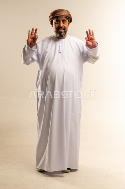 تعابير الرضا والسرور، الشعور بالاعجاب والقبول، بورتريه لرجل عربي خليجي عماني يرتدي الدشداشة والعمامة يقف باستقامة يرفع يديه للأعلى بإيماءات الجودة، صورة الجسم بالطول الكامل، خلفية بيضاء