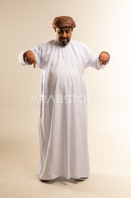 حركات يدين تدل على هذا المكان (هنا)، تحديد موقع شيء ما، بورتريه لرجل عربي خليجي عماني مبتسم يرتدي الدشداشة والعمامة يشير بأصبعي السبابة نحو الأسفل، صورة الجسم بالطول الكامل، خلفية بيضاء