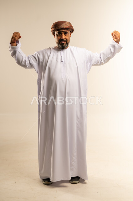 رفع اليدين لإظهار القوة، بورتريه لرجل عربي خليجي عماني يرتدي الدشداشة والعمامة يقوم بعرض عضلاته، النظر إلى الكاميرا بإيماءات الجدية، ممارسة الرياضة والحفاظ على جسم صحي، التمتع باللياقة وبنية جسدية قوية، صورة الجسم بالطول الكامل، خلفية بيضاء