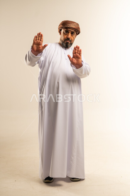 Expressions of refusal and abstention, order to stop doing something, portrait of an Arab Gulf Omani man wearing a dishdasha and turban raising his hands in a gesture of prohibition, feeling annoyed and angry, full body image, white background