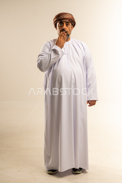 الشعور بالدهشة والصدمة والذهول, التفاجؤ من شيء ما، بورتريه لرجل عربي خليجي عماني يرتدي الدشداشة والعمامة يضع يديه على فمه بإيماءات التعجب، صورة الجسم بالطول الكامل، خلفية بيضاء