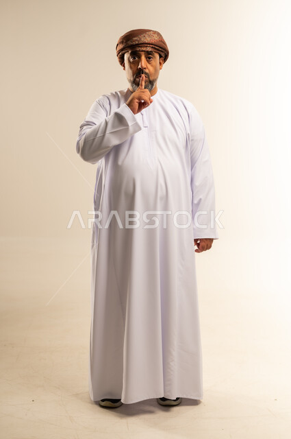Stop talking and order silence and quiet, portrait of an Arab Gulf Omani man wearing a dishdasha and turban raising his finger to his mouth with gestures asking for calm and not to disturb, full body image, white background