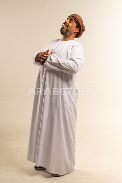 تعابير الضحك الهستيري، الشعور بالاطمئنان والراحة، بورتريه لرجل عربي خليجي عماني مبتسم يرتدي الدشداشة والعمامة يضع يديه على صدره بإيماءات الاستقبال والترحيب، صورة الجسم بالطول الكامل, خلفية بيضاء