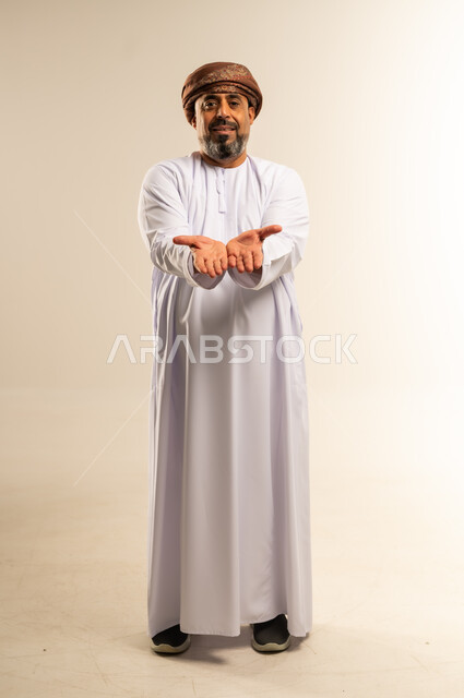 Presentation concept, looking at the camera with expressions of pleasure and happiness, portrait of a smiling Omani Gulf Arab man wearing a dishdasha and turban, opening his hands up with gestures of holding an item, full body photo, white background