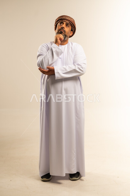 Finding a solution to a problem, contemplation and planning for the future, portrait of an Arab Gulf Omani man wearing a dishdasha and turban, putting his hand on his chin with gestures of thinking and concentration, expressions of absent-mindedness and contemplation, full body image, white background