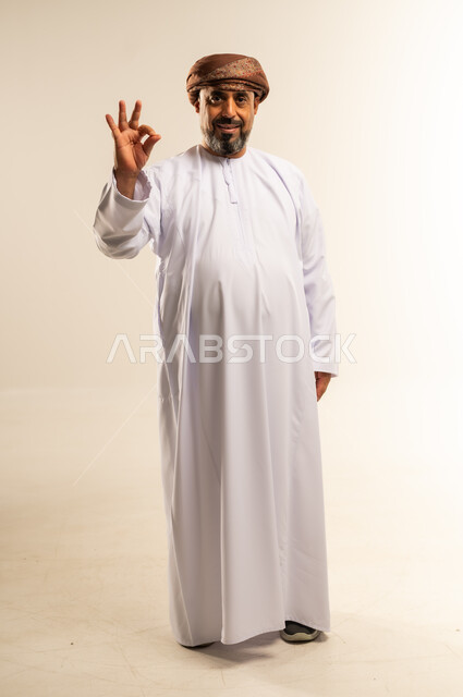 تعابير الرضا والسرور، الشعور بالاعجاب والقبول، بورتريه لرجل عربي خليجي عماني يرتدي الدشداشة والعمامة يقف باستقامة يرفع يده للأعلى بإيماءات الجودة، صورة الجسم بالطول الكامل، خلفية بيضاء