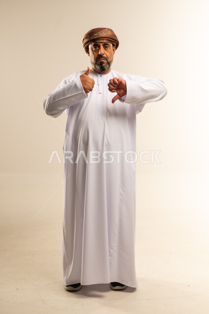 الحيرة والتردد بخصوص امر ما، بورتريه لرجل عربي خليجي عماني يرتدي الدشداشة والعمامة يرفع اصبع الابهام للاعلى والاسفل بإيماءات عدم الموافقه والموافقه، عدم الإعجاب والإعجاب، مفهوم الرفض والقبول، صورة الجسم بالطول الكامل, خلفية بيضاء