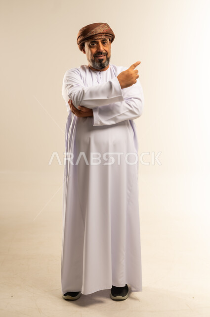 تحديد موقع شيء ما، حركات يدين تدل على هذا المكان (هنا)، بورتريه لرجل عربي خليجي عماني مبتسم يرتدي الدشداشة والعمامة يشير بأصبعي السبابة نحو اليسار، صورة الجسم بالطول الكامل، خلفية بيضاء