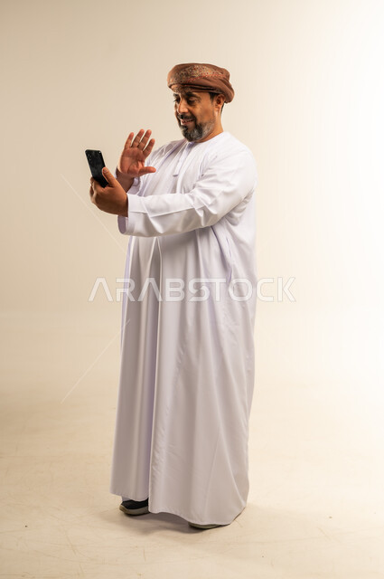 اجراء مكالمة فيديو، استخدام الأجهزة التقنية الحديثة، التواصل مع الأهل والأصدقاء، الاطمئنان على الأقارب وسماع اخبارهم، بورتريه من الجانب لرجل عربي خليجي عماني يرتدي الدشداشة والعمامة يحمل بيده الهاتف المحمول يرفع يده بايماءات السلام، صورة لكامل الجسم، خلفية بيضاء