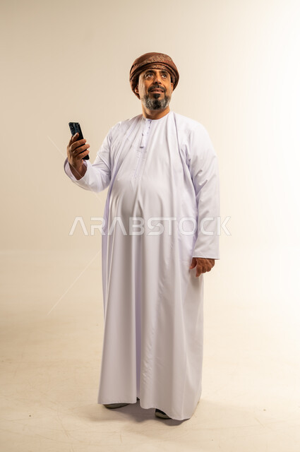 استخدام الأجهزة التقنية الحديثة، التواصل مع الأهل والأصدقاء، الاطمئنان على الأقارب وسماع اخبارهم، بورتريه لرجل عربي خليجي عماني يرتدي الدشداشة والعمامة يحمل بيده الهاتف المحمول بإيماءات السعادة، صورة الجسم بالطول الكامل، خلفية بيضاء
