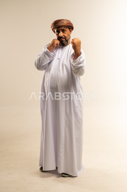 رفع اليدين لإظهار القوة، بورتريه لرجل عربي خليجي عماني يرتدي الدشداشة والعمامة يقوم بعرض عضلاته، النظر إلى الكاميرا بإيماءات الجدية، ممارسة الرياضة والحفاظ على جسم صحي، التمتع باللياقة وبنية جسدية قوية، صورة الجسم بالطول الكامل، خلفية بيضاء