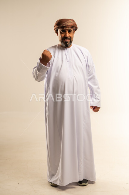 القوة والتحدي والحماس، مفهوم الانجاز وتحقيق الاهداف، بورتريه لرجل عربي خليجي عماني يرتدي الدشداشة والعمامة يرفع قبضة يده نحو الأعلى بإيماءات التشجيع، تعابير السرور والسعادة، صورة لكامل الجسم، خلفية بيضاء