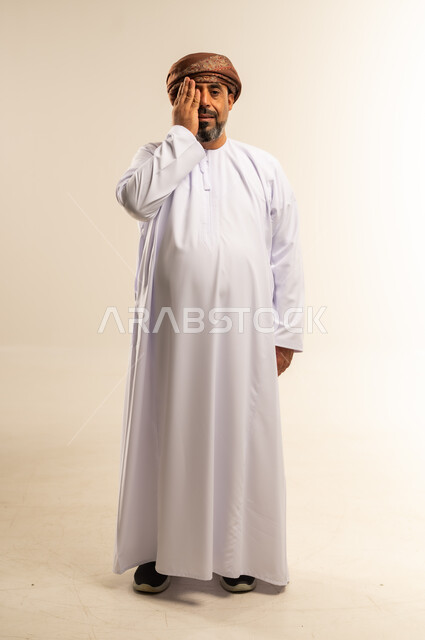 مشاكل عينيه وحالات مرضية، الحاجة للفحص والعلاج، مفهوم التهاب القرنية، بورتريه لرجل عربي خليجي عماني يرتدي الدشداشة والعمامة يضع يده على عينه بايماءات الألم، عدم القدرة على فتح العينين، صورة الجسم بالطول الكامل، خلفية بيضاء