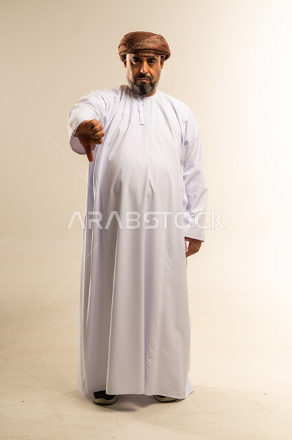 تعابير الرفض وعدم القبول، النظر إلى الكاميرا بإيماءات الانزعاج والسلبية، بورتريه لرجل عربي خليجي عماني يرتدي الدشداشة والعمامة يشير باصبع الابهام نحو الأسفل، صورة الجسم بالطول الكامل، خلفية بيضاء