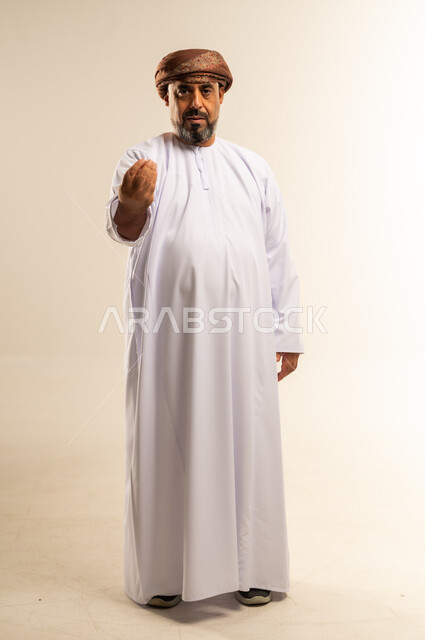 الهدوء وطلب الإنتظار، إمهال الوقت للقيام بشيء ما، حركة يد وإشارات، بورتريه لرجل عربي خليجي عماني يرتدي الدشداشة والعمامة يقف باستقامة يرفع يده بايماءات الصبر والتأن والتريث، صورة الجسم بالطول الكامل، خلفية بيضاء