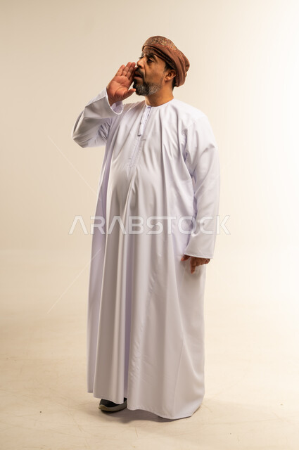 النداء بصوت منخفض والكلام بصوت عالي، مفهوم الدردشات والمحادثات، بورتريه لشاب عربي خليجي عماني مبتسم يرتدي الدشداشة و الكمه يضع يده على فمه بإيماءات المناداة، صورة الجسم بالطول الكامل، خلفية بيضاء