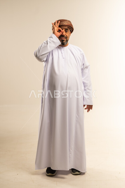 محاولة تقريب الأشياء البعيدة، التخطيط للأهداف والطموحات المستقبلية، حب الاكتشاف والمعرفة والمغامرة، التركيز على شيء ما، بورتريه لرجل عربي خليجي عماني يرتدي الدشداشة والعمامة يضع يده على عينيه بشكل عدسة بإيماءات البحث، صورة لكامل الجسم، خلفية بيضاء