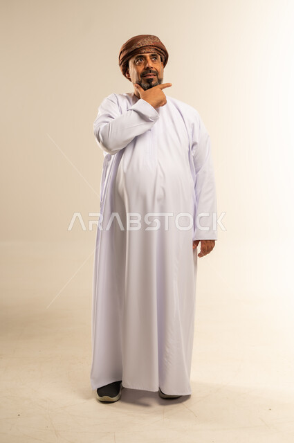 التأمل والتخطيط للمستقبل، بورتريه لرجل عربي خليجي عماني يرتدي الدشداشة والعمامة يضع يده على ذقنه بايماءات التفكير والتركيز، ايجاد حل لمشكلة ما، تعابير شرود الذهن والتمعن، صورة لكامل الجسم، خلفية بيضاء