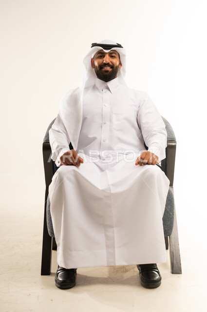 The concept of handsomeness, masculinity and strength, interest in elegance and external appearance, portrait of an Arab Gulf Qatari man wearing the traditional thobe and ghutra sitting on a comfortable chair looking at the camera with gestures of pleasure and self-confidence, white background