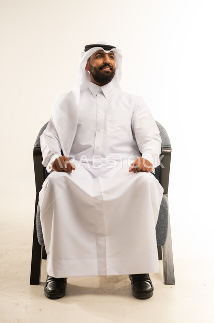 Concern for elegance and appearance, portrait of a Qatari Gulf Arab man wearing a traditional thobe and ghutra sitting on a comfortable chair looking up with gestures of pleasure and self-confidence, the concept of handsomeness, masculinity and strength, white background