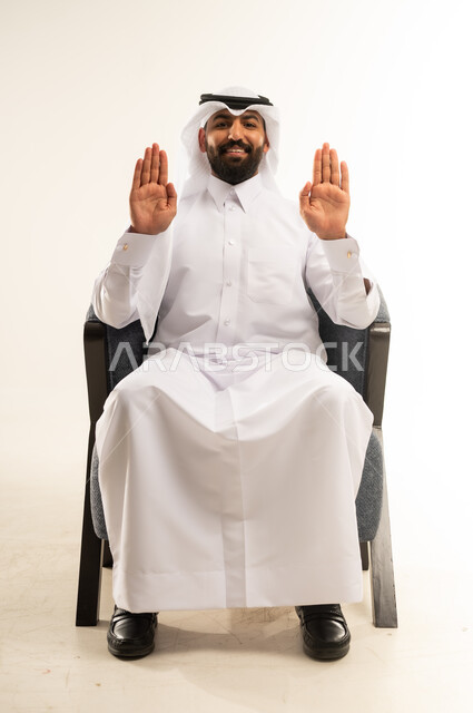مفهوم الاناقة والاهتمام بالمظهر الخارجي، عرض ظاهر الكف والنظر الى الكاميرا بتعابير الفرح والسرور، بورتريه لرجل عربي خليجي قطري مبتسم يرتدي الثوب والغترة يجلس على كرسي مريح يرفع كفيه مفتوحة للاعلى، خلفية بيضاء