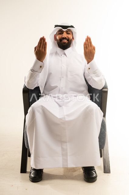 عرض باطن الكف والنظر الى الكاميرا بتعابير الفرح والسرور، بورتريه لرجل عربي خليجي قطري مبتسم يرتدي الثوب والغترة يجلس على كرسي مريح يرفع كفيه مفتوحة للاعلى، خلفية بيضاء