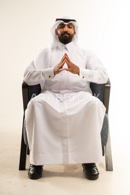 مفهوم الاناقة والاهتمام بالمظهر الخارجي, ايماءات الشعور بالحيرة وعدم التأكد،  بورتريه لرجل عربي خليجي قطري يرتدي الثوب التقليدي والغترة يجلس على كرسي مريح ينظر الى الكاميرا بتعابير السرور، خلفية بيضاء