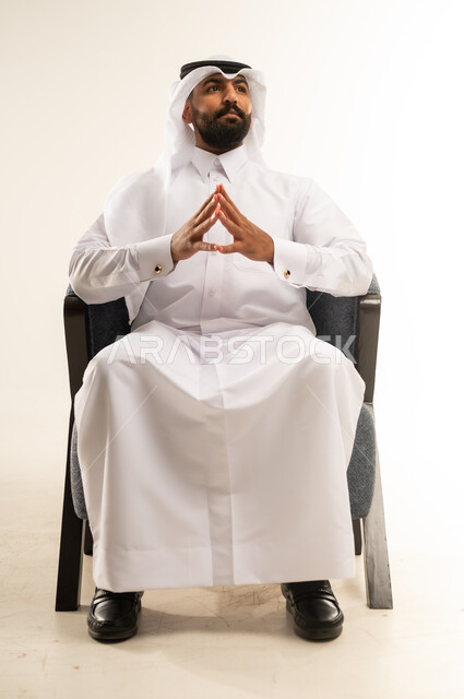الشعور بالحيرة وعدم التأكد, مفهوم الاناقة والاهتمام بالمظهر الخارجي، إيماءات التأمل والتمعن والتركيز, بورتريه لرجل عربي خليجي قطري يرتدي الثوب التقليدي والغترة يجلس على كرسي ينظر الى مكان ما بتعابير التفكير، التخطيط لحل مشكلة ما، خلفية بيضاء