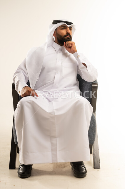 الشعور بالحيرة وعدم التأكد, مفهوم الاناقة والاهتمام بالمظهر الخارجي، إيماءات التأمل والتمعن والتركيز, بورتريه لرجل عربي خليجي قطري يرتدي الثوب التقليدي والغترة يجلس على كرسي يضع يده خلف ذقنه ينظر الى مكان ما بتعابير التفكير، التخطيط لحل مشكلة ما، خلفية بيضاء