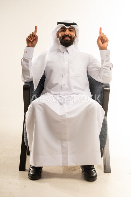 اشارة رقم واحد، النظر إلى الكاميرا بإيماءات الفرح والسرور، بورتريه لشاب عربي خليجي قطري مبتسم يرتدي الثوب التقليدي والغترة يجلس على كرسي مريح يشير بإصبعه السبابة للاعلى  إلى شيء محدد بتعابير هذا المكان هنا، خلفية بيضاء .