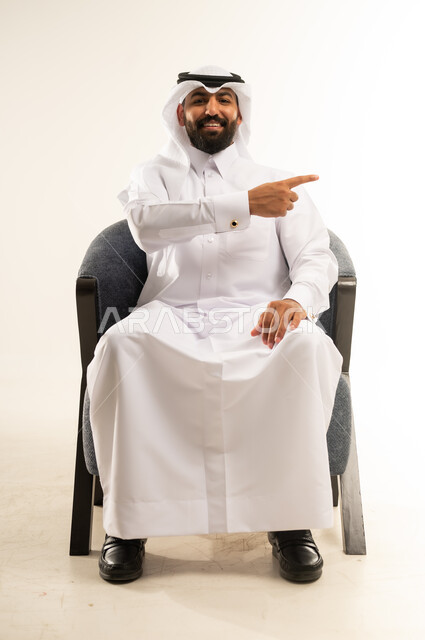 النظر إلى الكاميرا بإيماءات الفرح والسرور، بورتريه لشاب عربي خليجي قطري مبتسم يرتدي الثوب التقليدي والغترة يجلس على كرسي مريح يشير بإصبعه السبابة جانبا  إلى شيء محدد بتعابير هذا المكان هنا، خلفية بيضاء .