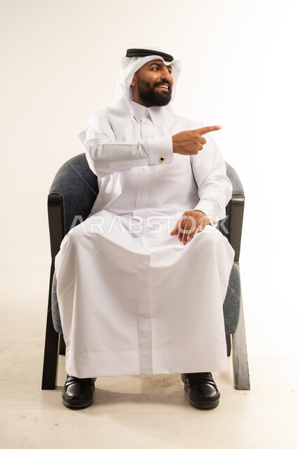 النظر إلى شيء ما جانبا بايماءات السرورر، بورتريه لشاب عربي خليجي قطري مبتسم يرتدي الثوب التقليدي والغترة يجلس على كرسي مريح يشير بإصبعه السبابة إلى شيء محدد بتعابير هذا المكان هنا، خلفية بيضاء .