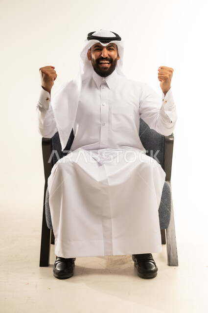 مفهوم الانجاز وتحقيق الاهداف، بورتريه لرجل عربي خليجي قطري مبتسم يرتدي الثوب التقليدي والغترة يجلس على كرسي مريح يرفع قبضة يديه للاعلى بإيماءات الفوز والانتصار والنجاح, النظر الى الكاميرا بتعابير السعادة والقوة والسرور، خلفية بيضاء