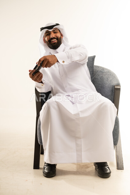 قضاء اوقات ممتعة بألعاب الفيديو الترفيهية في عطلة نهاية الاسبوع، بورتريه لرجل عربي خليجي قطري مبتسم يرتدي الثوب والغترة يجلس على كرسي مريح يمسك بيده عصا التحكم بتعابير الاندماج والسرور، اللعب والاستمتاع بأوقات الفراغ، دمج التقنية والتكنولوجيا في الحياة اليومية، خلفية بيضاء