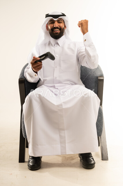 مفهوم الانجاز وتحقيق الاهداف، الاندماج والتفاعل في ألعاب الفيديو الترفيهية، بورتريه لرجل عربي خليجي قطري مبتسم يرتدي الثوب والغترة يجلس على كرسي مريح يمسك بيده عصا التحكم يقوم برفع قبضة يده بتعابير الفوز والنجاح، دمج التقنية والتكنولوجيا في الحياة اليومية، خلفية بيضاء