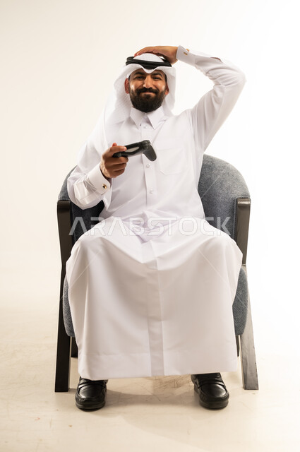 الاندماج والتفاعل في ألعاب الفيديو الترفيهية، بورتريه لرجل عربي خليجي قطري مبتسم يرتدي الثوب والغترة يجلس على كرسي مريح يمسك عصا التحكم يقوم بوضع يده عبى رأسه بتعابير الخسارة والهزيمة والانزعاج ، دمج التقنية والتكنولوجيا في الحياة اليومية، خلفية بيضاء