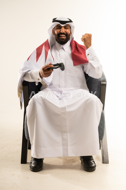 مفهوم الانجاز وتحقيق الاهداف، حب الوطن والانتماء اليه، الاندماج والتفاعل في ألعاب الفيديو الترفيهية، بورتريه لرجل عربي خليجي قطري مبتسم يرتدي الثوب والغترة وعلم دولة قطر يجلس على كرسي مريح يمسك بيده عصا التحكم يقوم برفع قبضة يده بتعابير الفوز والنجاح والفخر، خلفية بيضاء