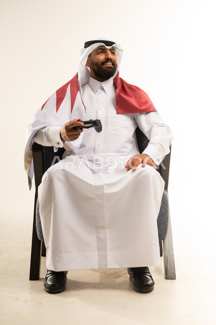 الاندماج والتفاعل في ألعاب الفيديو الترفيهية، مفهوم الانجاز وتحقيق الاهداف، حب الوطن والانتماء اليه، بورتريه لرجل عربي خليجي قطري مبتسم يرتدي الثوب والغترة وعلم دولة قطر يجلس على كرسي مريح يمسك بيده عصا التحكم ينظر الى مكان ما بتعابير الفرح والسرور، خلفية بيضاء