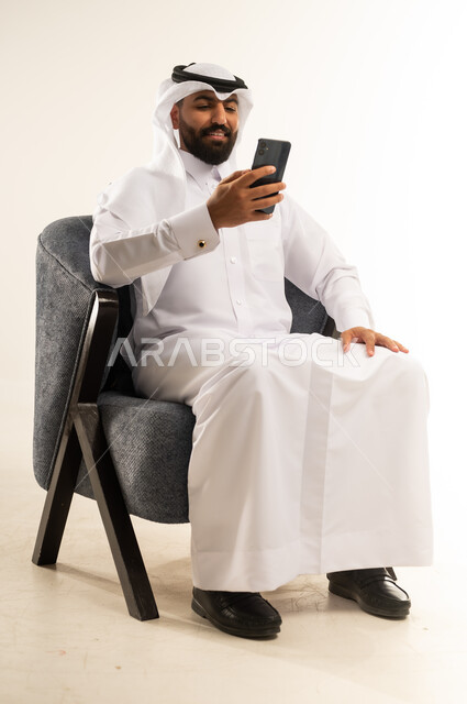دمج التكنولوجيا في الحياة اليومية، استخدام التقنيات والأجهزة الحديثة، تصفح التطبيقات ومواقع التواصل الاجتماعي عبر الانترنت، دردشة ومحادثات وتعارف، بورتريه لرجل عربي خليجي قطري مبتسم يرتدي الثوب التقليدي والغترة يجلس على كرسي مريح يقوم باستخدام الهاتف المحمول، خلفية بيضاء