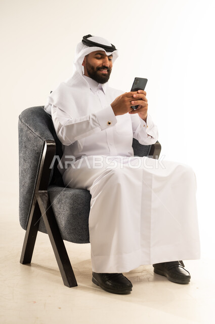 دمج التكنولوجيا في الحياة اليومية، استخدام التقنيات والأجهزة الحديثة، تصفح التطبيقات ومواقع التواصل الاجتماعي عبر الانترنت، دردشة ومحادثات وتعارف، بورتريه لرجل عربي خليجي قطري مبتسم يرتدي الثوب التقليدي والغترة يجلس على كرسي مريح يقوم باستخدام الهاتف المحمول، خلفية بيضاء