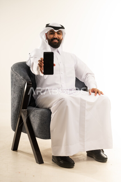Taking advantage of mobile services and applications, integrating technology and modern devices into daily life, portrait of a smiling Qatari Gulf Arab man wearing traditional thobe and ghutra sitting on a comfortable chair displaying a blank black screen on a mobile phone, looking at the camera with expressions of joy and happiness, white background