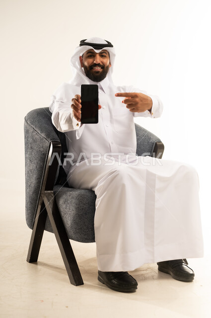 Benefiting from mobile services and applications, integrating technology and modern devices into daily life, portrait of a smiling Qatari Gulf Arab man wearing a thobe and ghutra sitting on a comfortable chair displaying a blank black screen on a mobile phone, pointing with his index finger to the device with expressions of joy and happiness, white background