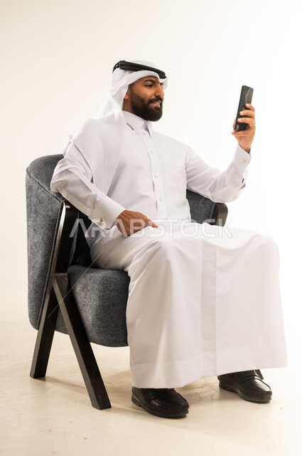 دمج التكنولوجيا في الحياة اليومية، استخدام التقنيات والأجهزة الحديثة، تصفح التطبيقات ومواقع التواصل الاجتماعي عبر الانترنت، دردشة ومحادثات وتعارف، بورتريه لرجل عربي خليجي قطري مبتسم يرتدي الثوب التقليدي والغترة يجلس على كرسي مريح يقوم باستخدام الهاتف المحمول، خلفية بيضاء