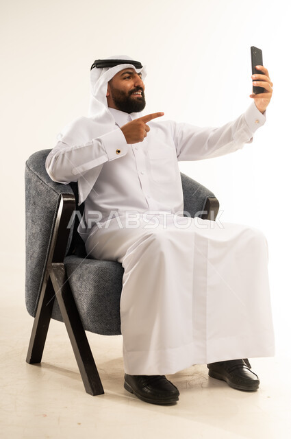 دمج التكنولوجيا والاجهزة الحديثة في الحياة اليومية، بورتريه لرجل عربي خليجي قطري مبتسم يرتدي الثوب التقليدي والغترة يجلس على كرسي مريح يقوم بإلتقاط صور سيلفي تذكارية باستخدام الهاتف المحمول، التواصل مع الاهل والاصدقاء، خلفية بيضاء